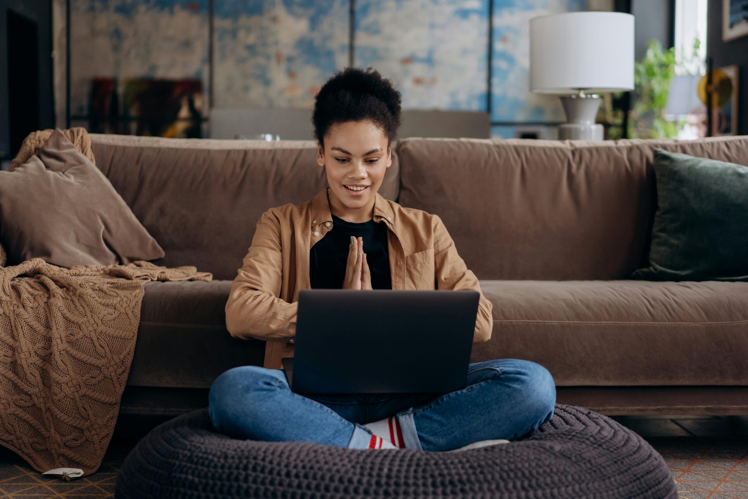 Imagem horizontal, com uma mulher negra (parda) no centro, sentada com pernas cruzadas e um notebook preto no colo. Atrás dela há um sofá marrom ocupando a largura da imagem. Atrás do sofá, no quadrante superior direito há um abajour branco. Predomina na imagem tons terrosos.