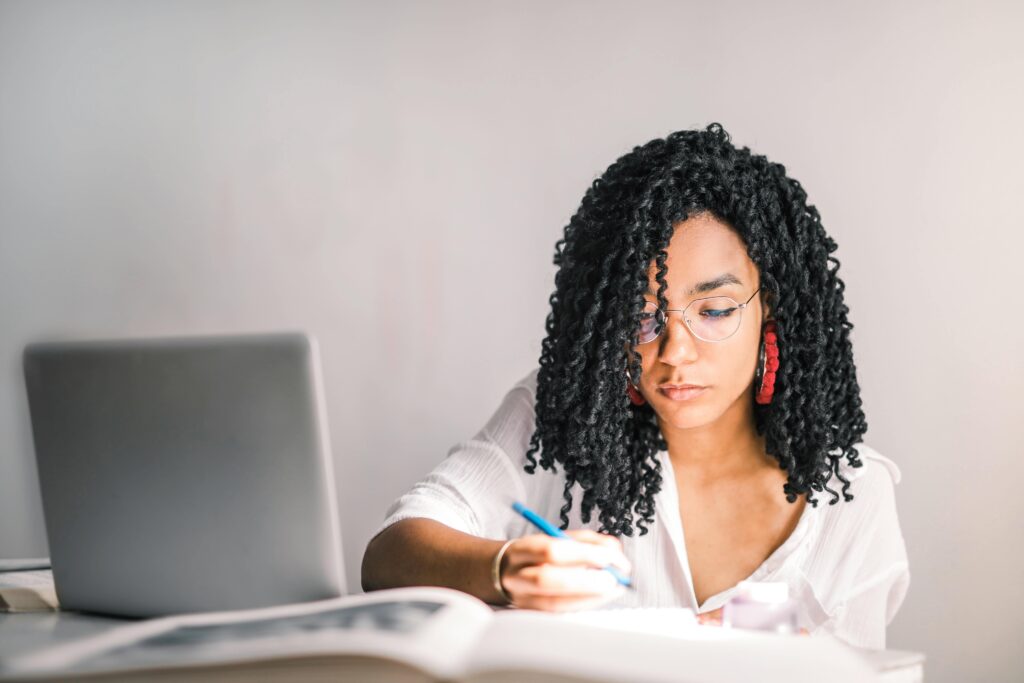 Imagem horizontal. No lado direito, há uma jovem negra (parda) com cabelo cacheado preto e brincos de argola vermelho. Ela veste uma blusa branca e essa cor é predominante na imagem porque é a cor da parede de fundo, da mesa em que ela escreve e nos livros de consulta. No lado esquerdo da imagem, há um notebook cinza.